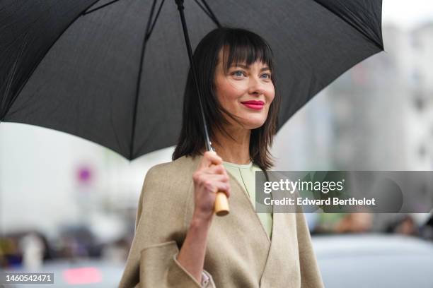 Guest wears a black umbrella, a pale green long silk dress, a beige wool with brown silk large belt long coat from Fendi, outside Fendi, during Paris...