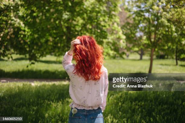 young woman with long curly red hair enjoys sunny weather in the spring. warm spring day. green grass lawn in the park. - dyed red hair fotografías e imágenes de stock