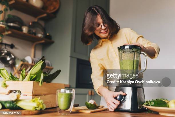 healthy eating, cooking, vegetarian food, dieting and people concept - close up of young woman with blender and green vegetables making detox shake or smoothie at home - mixer stock-fotos und bilder