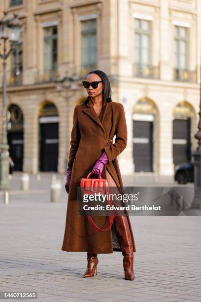 Emilie Joseph wears black sunglasses, a brown wool long trench coat, purple shiny leather gloves, a red shiny leather handbag, brown shiny leather...