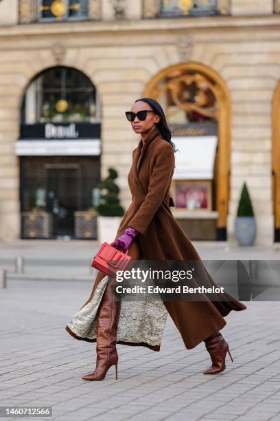 Emilie Joseph wears black sunglasses, a brown wool long trench coat, purple shiny leather gloves, a red shiny leather handbag, brown shiny leather...