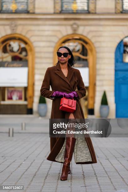 Emilie Joseph wears black sunglasses, a brown wool long trench coat, purple shiny leather gloves, a red shiny leather handbag, brown shiny leather...