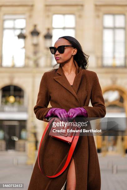 Emilie Joseph wears black sunglasses, a brown wool long trench coat, purple shiny leather gloves, a red shiny leather handbag, during a street style...