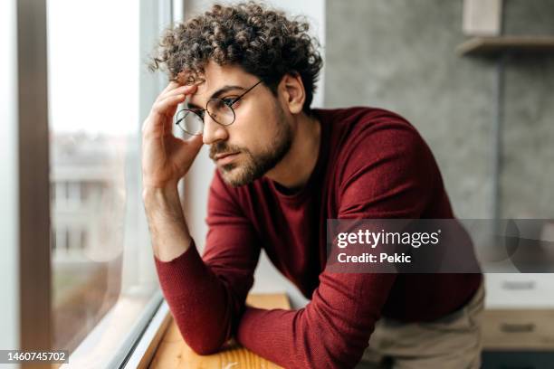 porträt eines depressiven mannes, der über seine gedanken nachdenkt, während er neben dem fenster in der wohnung steht - headache stock-fotos und bilder