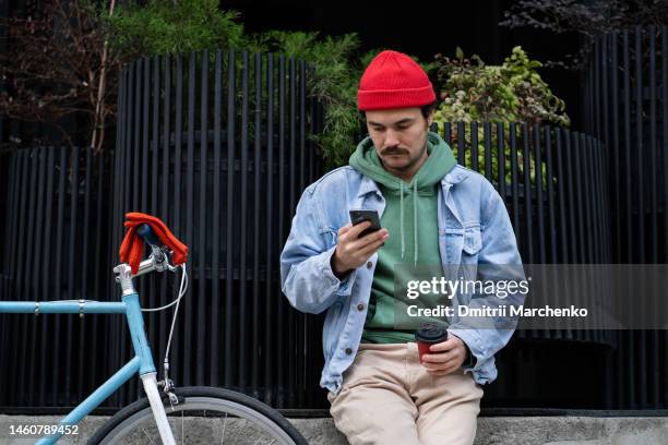 sociable young man watching mobile phone answering message to friends sits on street near bike - mid adult men stock pictures, royalty-free photos & images