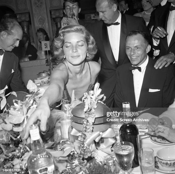 Lauren Bacall reaching for Smirnoff Vodka bottle on table as Frank Sinatra smiles looking on at an after-theater party following the world premiere...