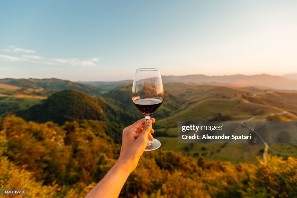 Man holding a glass of red wine among hills and mountains, personal perspective view