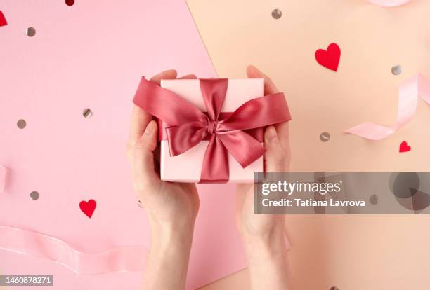 female hands holding gift box with satin magenta ribbon on festive pink background with confetti and hearts. valentines day, international women day, mothers day concept - valentine card stock pictures, royalty-free photos & images