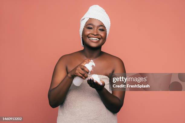 a happy beautiful woman wrapped in a towel looking at camera while holding a spray bottle - spray foam stock pictures, royalty-free photos & images