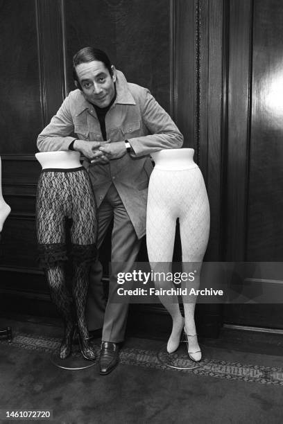 Portrait of American costume jewelry designer Kenneth Jay Lane leaning on a pair of mannequins' legs in textured stockings.