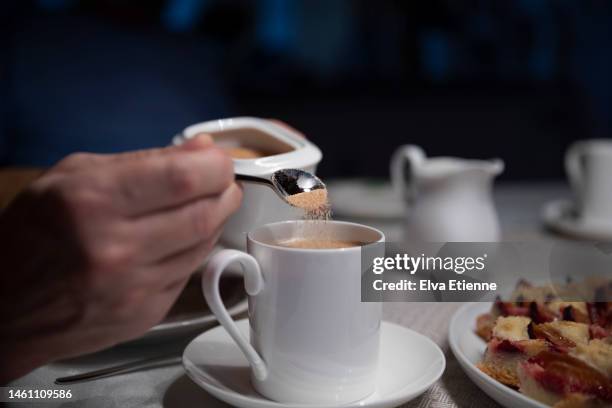 close-up of brown sugar being added to a cup of coffee by a man at a dining table with white porcelain crockery and sweet food on a plate. - brown sugar stock pictures, royalty-free photos & images