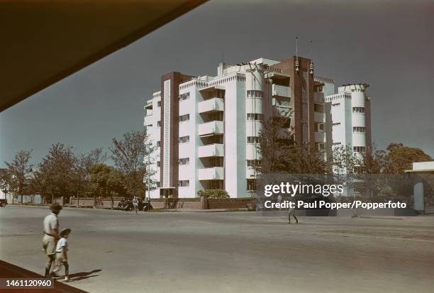 Pedestrians walk across a road past cars parked in front of an Art Deco styled apartment block building in the city of Bulawayo in Southern Rhodesia...