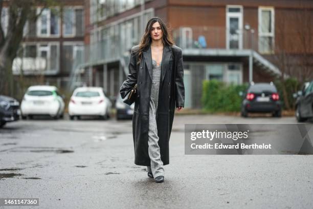 Ilirida Krasniqi wears silver earrings, a gray wool heart neck / shoulder-off /large jumpsuit, a black shiny leather oversized long coat, a black...