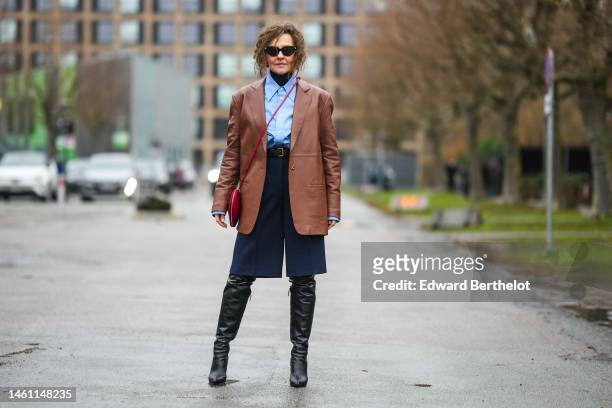 Renia Jaz wears black sunglasses, a black wool turtleneck pullover, a pale blue shirt, a brown shiny leather oversized blazer jacket, a black shiny...