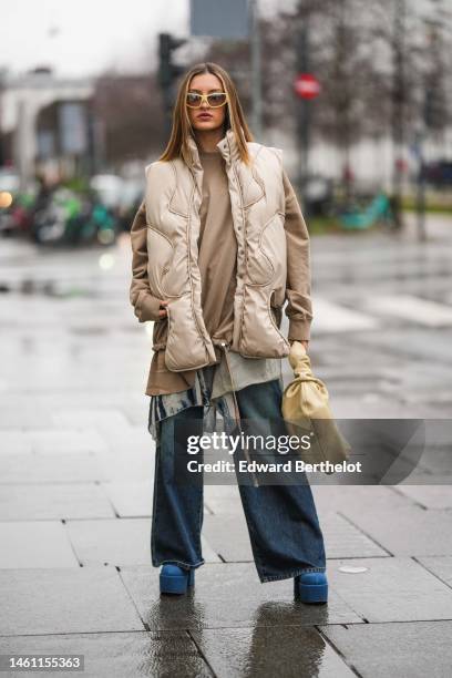 Guest wears pale yellow sunglasses, a beige oversized sweater, a beige quilted pattern sleeveless / oversized puffer jacket, a beige matte leather...