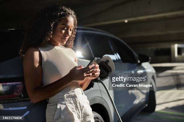 woman using smart phone by electric car at parking garage - red car wire fotografías e imágenes de stock