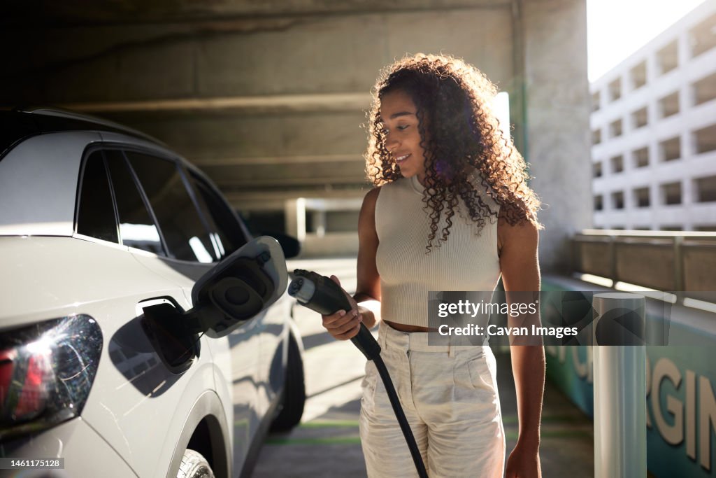 Young woman holding electric plug by car at charging station