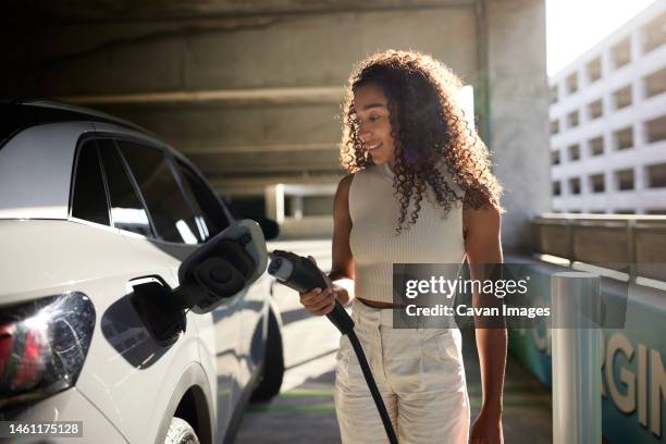 young woman holding electric plug by car at charging station - electrical plug foto e immagini stock