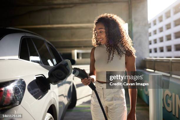young woman holding electric plug by car at charging station - elektrische auto stockfoto's en -beelden