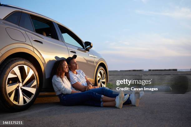 smiling woman sitting with boyfriend against car during sunset - sustainable transportation stock pictures, royalty-free photos & images