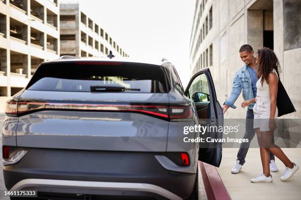 man opening door of electric car for girlfriend standing on footpath - gallanterie stock-fotos und bilder