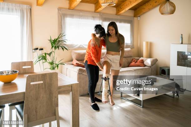 woman helping daughter with broken leg to move from sofa - orthopaedic equipment stock-fotos und bilder