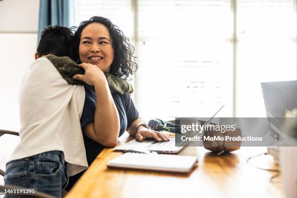 woman working from home is interrupted by her son at dining room table - family hugging bright stockfoto's en -beelden