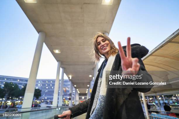 low angle view of stylish cheerful woman waiting on moving walkway in airport terminal looking at camera carrying travel suitcase gesturing victory peace symbol with fingers - palm tree leaf stock pictures, royalty-free photos & images