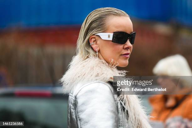Guest wears black and white sunglasses, silver earrings, a silver shiny leather with white fur interior and borders coat, outside Aeron , during the...
