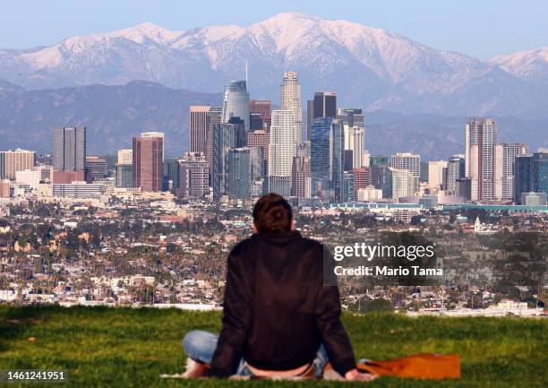 Person sits on a hillside with a view of the downtown skyline, with the snow-capped San Gabriel Mountains visible beyond, on January 31, 2023 in Los...