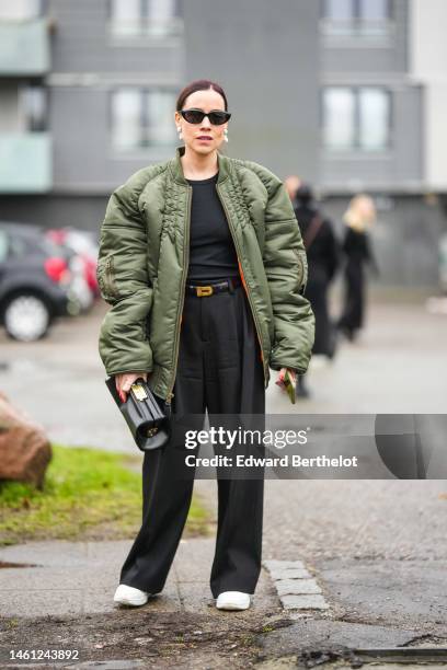 Guest wears black sunglasses, a black t-shirt, a khaki nylon bomber coat, a black shiny leather handbag, black large suit pants, a black shiny...