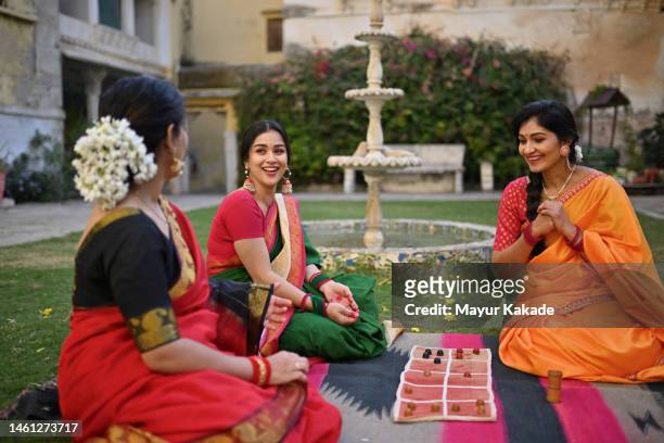 beautiful indian women playing vintage board game sitting on a lawn in a courtyard - woman in red sari stock pictures, royalty-free photos & images