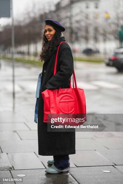 Guest wears a navy blue felt wool beret, a black long wool coat, a red oversized shiny leather Telfar shoulder bag, navy blue denim large pants, pale...