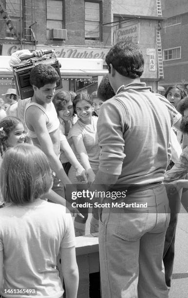 View, from behind, of an man leading a game of Three-card Monte as onlookers, one holding a portable radio on his shoulder, point at the cards on a...