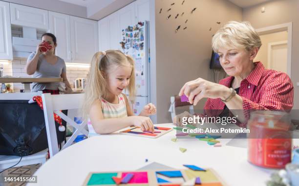 piccola, sorridente bambina di cinque anni che gioca con il puzzle tangram al tavolo della cucina con sua nonna, la madre sta dietro di loro, bevendo il tè. - "alex potemkin" or "krakozawr" foto e immagini stock