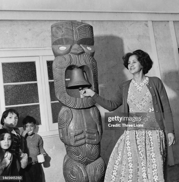 Teacher rings a bell housed in a wooden figure, carved by Māori artisans for a school in New Zealand, circa 1955.