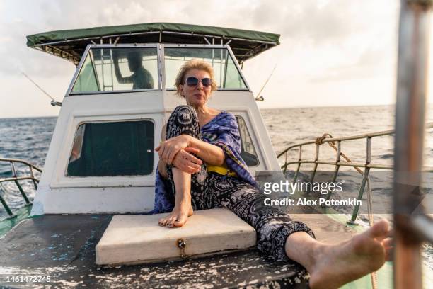 una donna europea attiva di 50 anni, una turista, sta riposando sul ponte di una piccola barca da pesca durante la battuta di pesca d'altura in sri lanka quando un capitano sta governando la nave da una tuga sullo sfondo. - "alex potemkin" or "krakozawr" foto e immagini stock