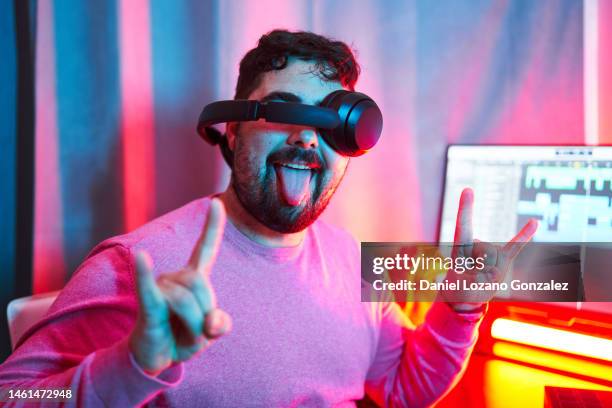 fun portrait of man with a wireless headphones bad located on his head covering his eyes and looking at camera while making the sign of horns with his hands. color led lighting. - sound engineer stock pictures, royalty-free photos & images