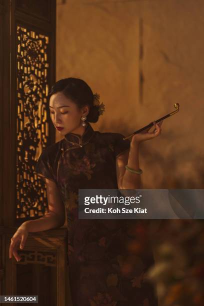 ritratto di una bella donna cinese in qi pao che tiene la sua sigaretta mentre si appoggia a un divisorio di legno fotografato in un ambiente di ritratto in studio - abito tradizionale cinese foto e immagini stock