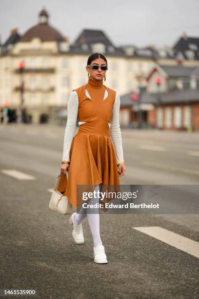 Melanie Darmon wears white and black print pattern sunglasses from Balenciaga, a white long sleeves t-shirt, a brown camel shiny leather turtleneck /...