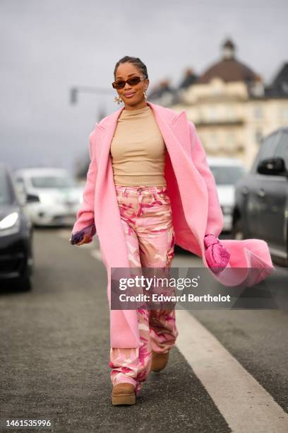 Ellie Delphine wears brown sunglasses from Miu Mui, gold earrings, a beige ribbed t-shirt, a pale pink wool oversized long coat, a pink shiny leather...