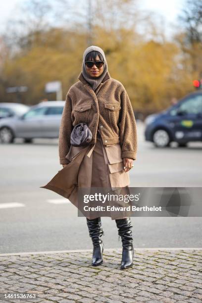 Loicka Grace wears black sunglasses, a pale brown wool hoodie, a brown fluffy buttoned jacket, a brown long pockets details knees dress, a brown...
