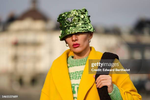 Katya Tolstova wears a green large sequined bob hat from Prada, silver earrings, a green and white latte wool print pattern pullover, a yellow wool...