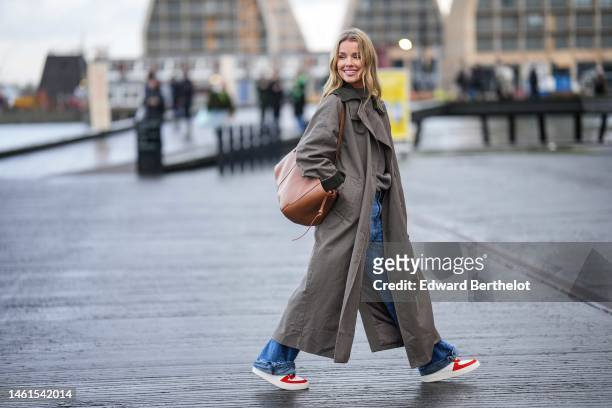Guest wears a gray brown ribbed wool high neck pullover, a brown long trench coat, a camel shiny leather shoulder bag, blue denim ripped wide legs...