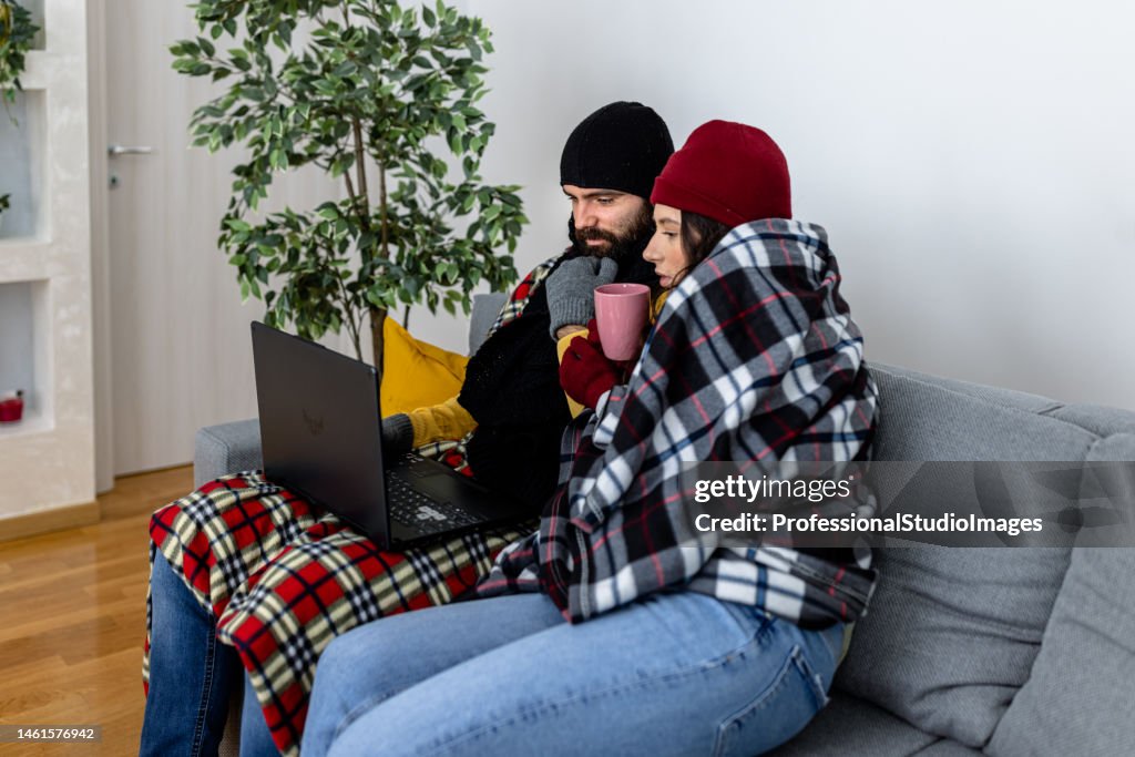 A man and woman in warm clothing are shivering and wearing a blanket and freezing at home while warming up with hot tea.