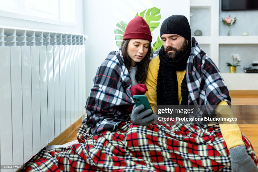 A young couple is suffering from a cold temperature during an energetic crisis and trying to warm up near the radiator.