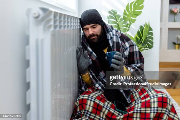 un homme portant des vêtements chauds gèle à la maison et essaie de se réchauffer avec du thé assis près du radiateur. - lowering photos et images de collection