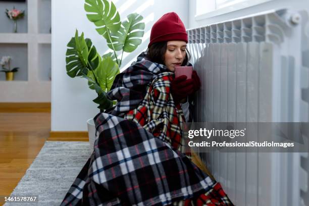 a woman wearing warm clothes is freezing at home and trying to warm up with tea sitting by the radiator. - broken air conditioner stock pictures, royalty-free photos & images