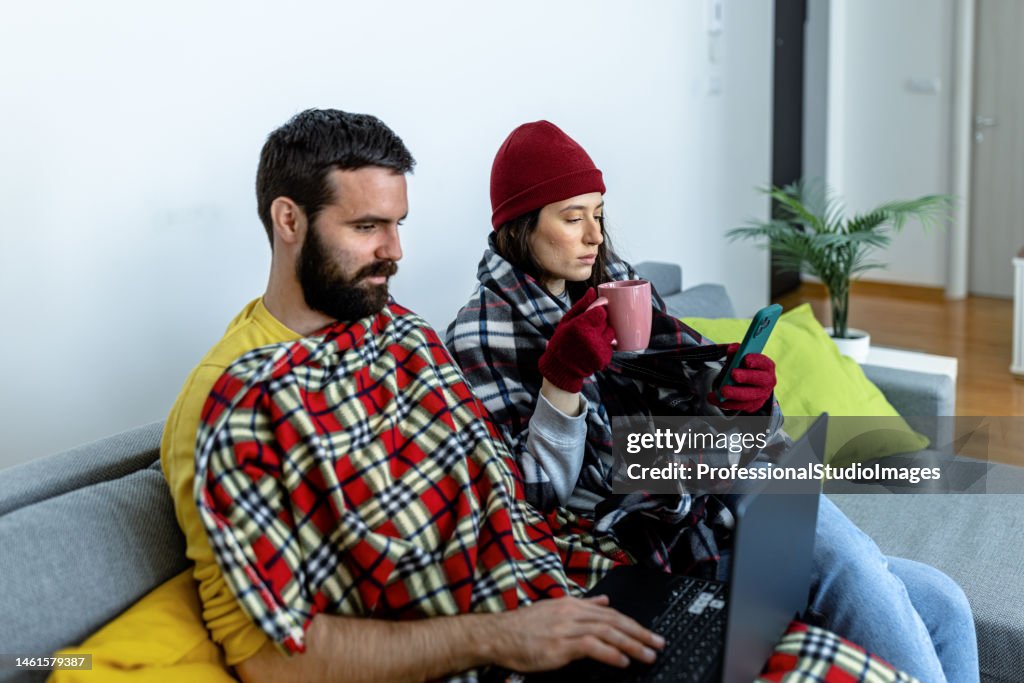 A man and woman wearing warm clothes are freezing at home and trying to warm up by sitting under a blanket on a sofa and using a laptop and phone.