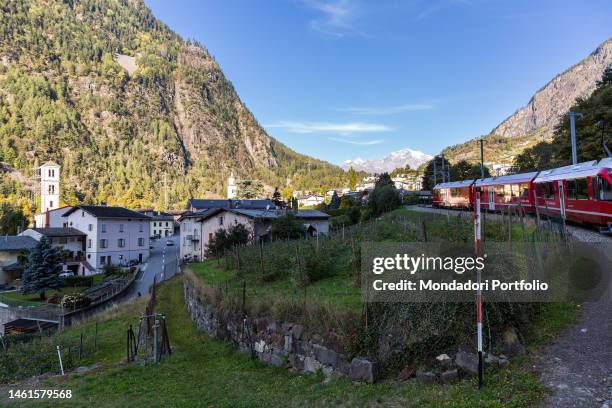 The passage of the Red Train of Bernina in the railway section located near the center of Brusio. In the background, the bell tower of the church of...